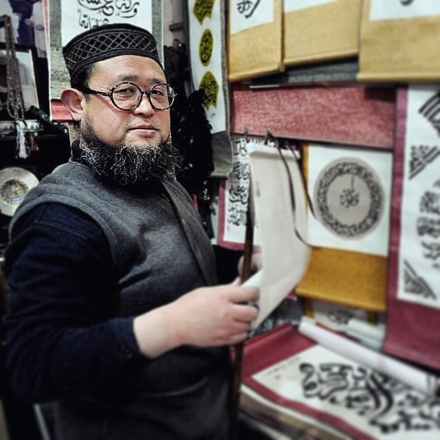 a Chinese Muslim man in a traditional cap standing in front of a wall hung with various scrolls with Arabic writing on them