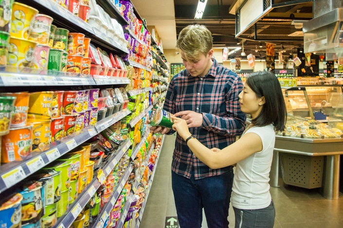a blond man and a Chinese woman looking at some Chinese instant noodles in a supermarket