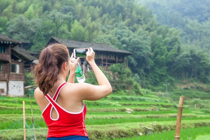 a brown haired girl with her back to the camera takes a picture of a rural scene in China on her phone