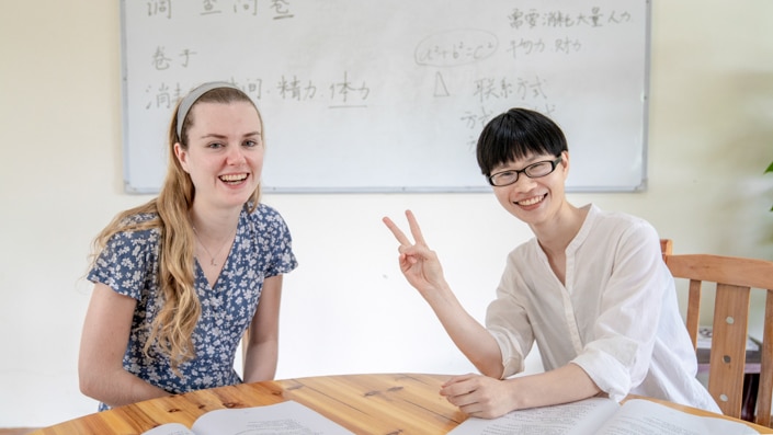 a blond student with her Chinese teacher sitting at a table in front of a whiteboard