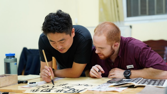 a red haired man watching a Chinese man write Chinese characters with a calligraphy brush