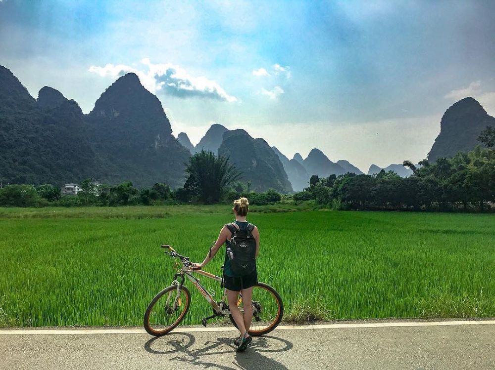 a blond woman serving as her own Yangshuo travel guide stands with a bike in front of a rice field flanked by karst peaks