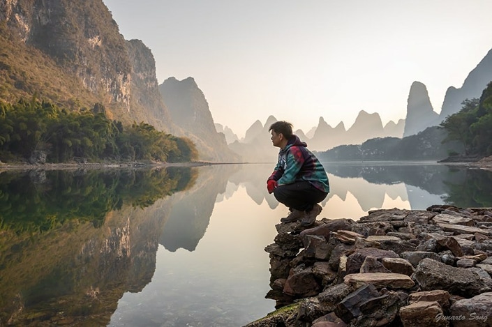 a hiker serving as his own Yangshuo travel guide squats beside a river with karst mountains in the background