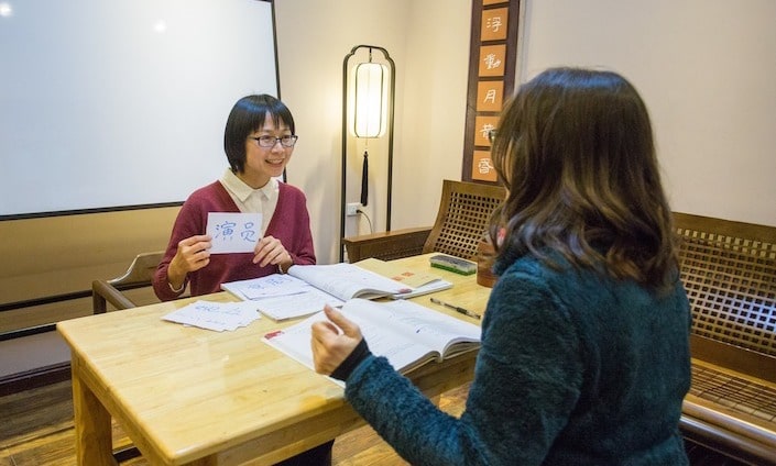 a short haired Chinese woman holding up a flashcard with a Chinese character on it for a female western student seated across from her at a table