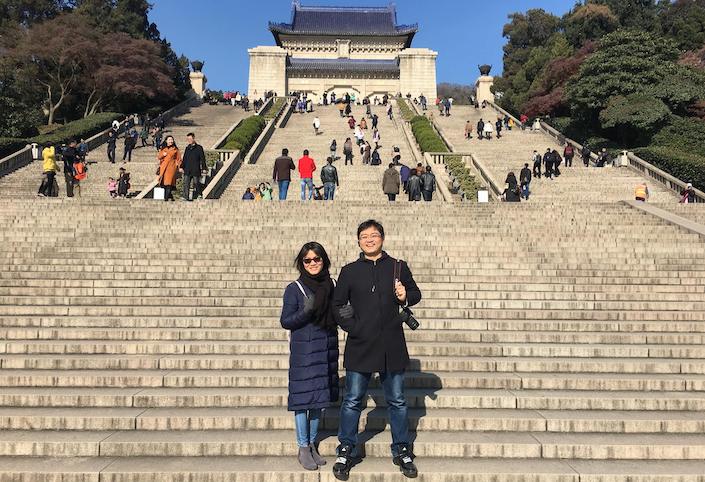 a man and a woman pose on the steps in front of the Sun Yatsen Mausoleum in Nanjing while other tourists climb the stairs in the background above them
