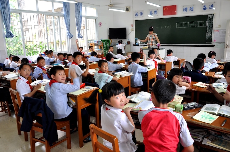 a photo taken from the back of a room showing a classroom full of young Chinese students with a teacher standing in front of a blackboard facing the class