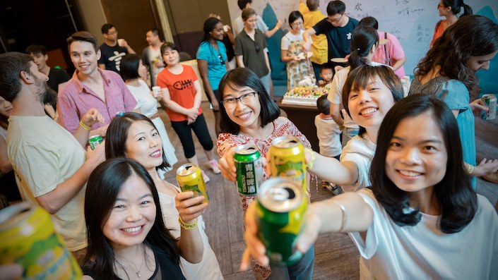 a group of young Chinese women toasting the camera with a canned beverage in their hands while multiple other people talk in the background