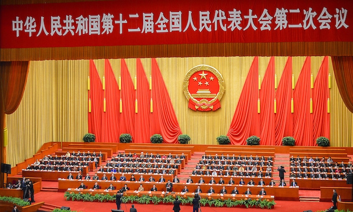 photo of a meeting of Chinese government officials with the seal of the People's Republic and two rows of red flags against a yellow wall behind them and a red banner with white Chinese characters above