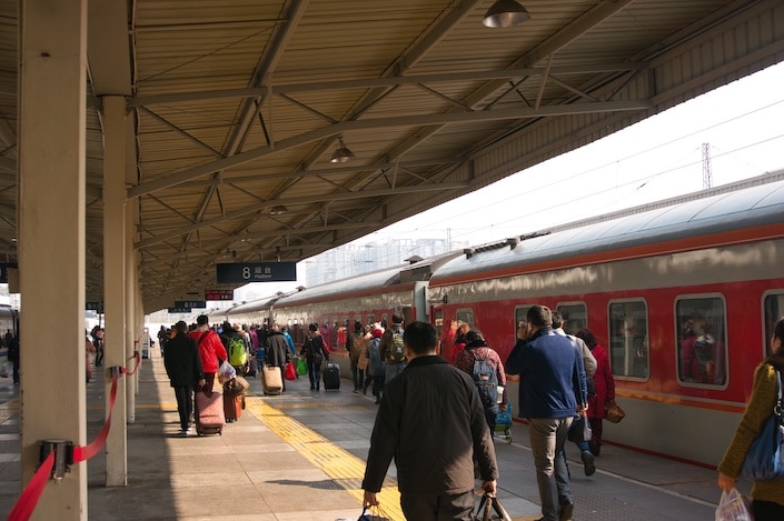 people carrying luggage walking on a covered platform next to an old-fashioned looking red train in China