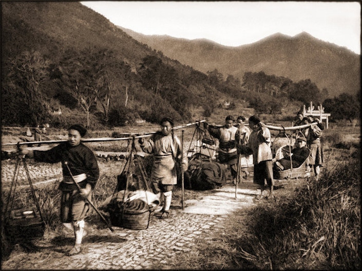 an old black and white photograph of farmers with shoulder poles walking along a path in the countryside with low mountains in the background