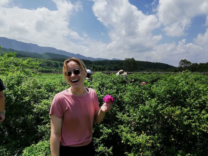 a woman wearing sunglasses and a pink T-shirt holding a flower in a field with mountains in the background on a sunny day