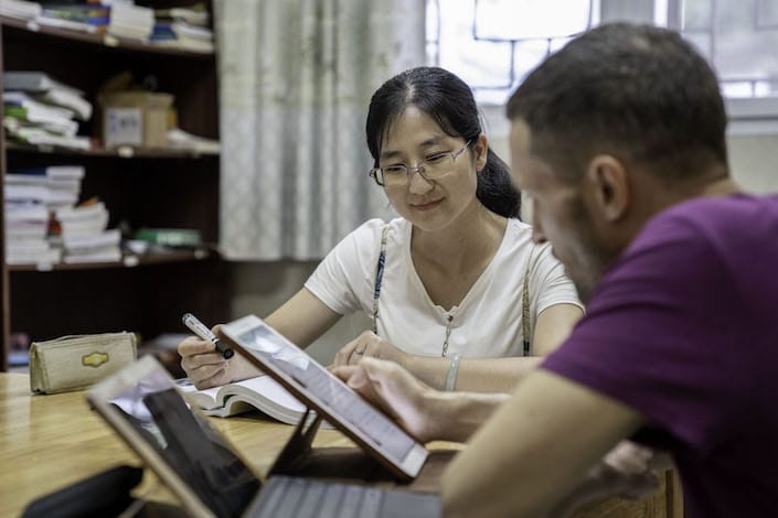 a female Chinese teacher looks on as a western man in a purple T-shirt uses a tablet to study Chinese