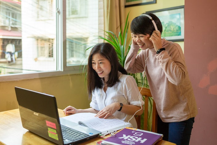 Two Chinese women, one seated and one standing with headphones, look at a laptop computer on a table