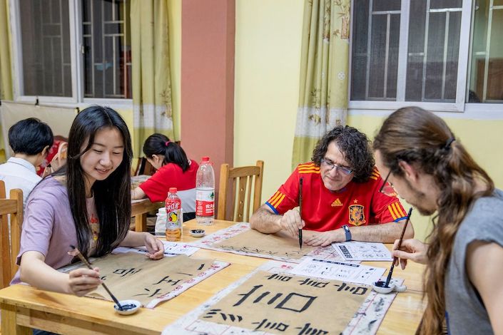 a Chinese woman and two western men with long hair sit at a table practicing Chinese calligraphy