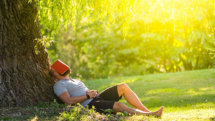 a man sitting in the grass outdoors while leaning against a tree with a red book covering his face