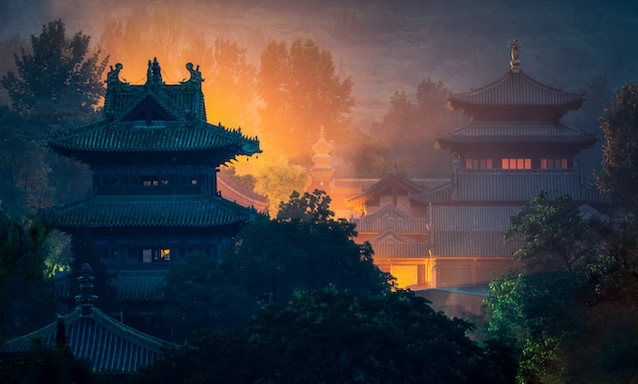 two buildings of the Shaolin Temple complex surrounded by trees and lit up with an orange glow
