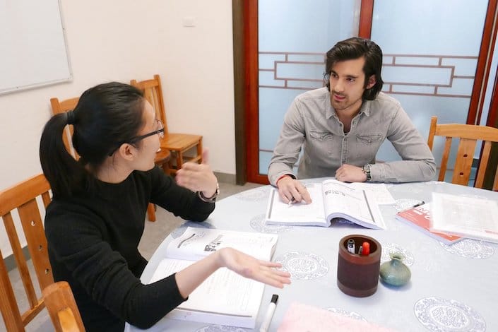 a western man with long hair having class with a female Chinese teacher while sitting around a circular table with several textbooks open on it