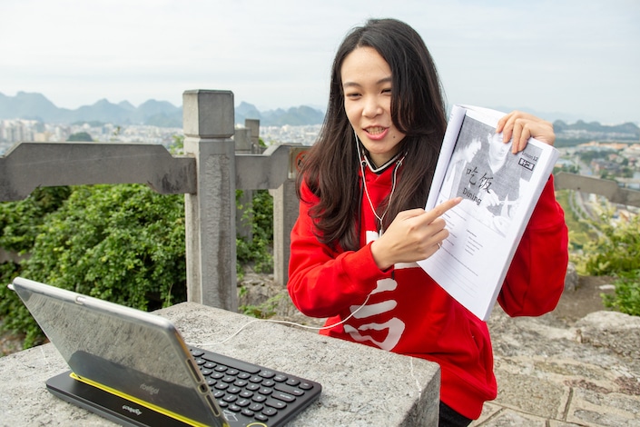 chinese teacher sitting outdoors holding textbook toward her laptop to show her online student something in the book