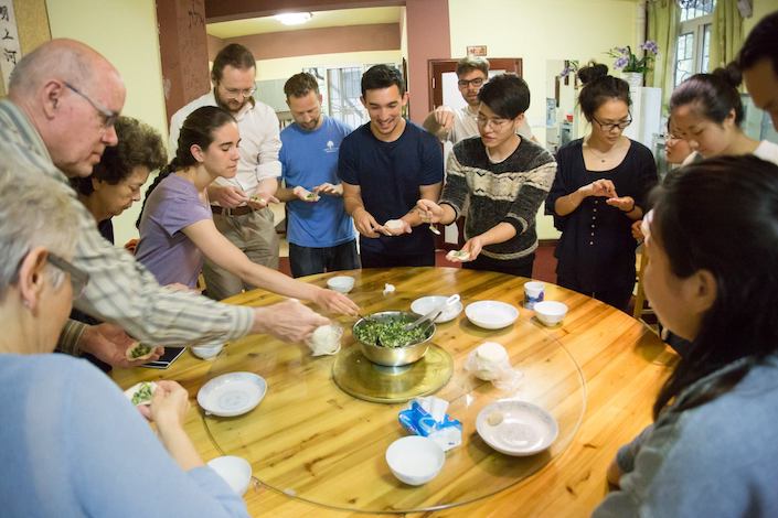 a group of young people standing around a round wooden table making chinese dumplings