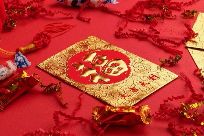a gold and red hongbao with the Chinese character for "luck" written on it laying on a red surface with candy scattered around it