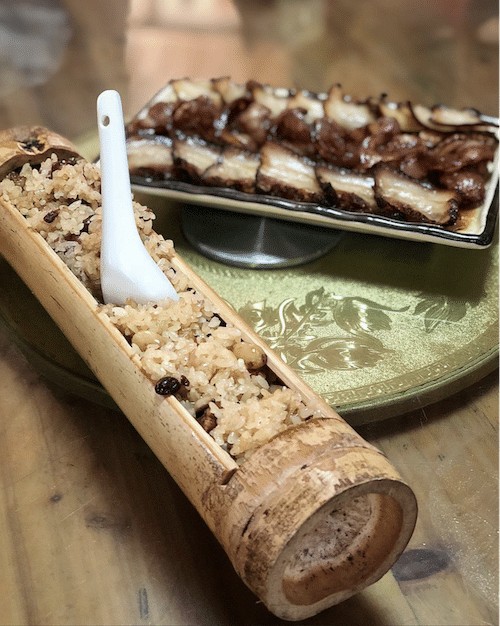 a bamboo tube stuffed with rice with a porcelain Chinese spook stuck in it next to a plate of cured meat on a wooden table