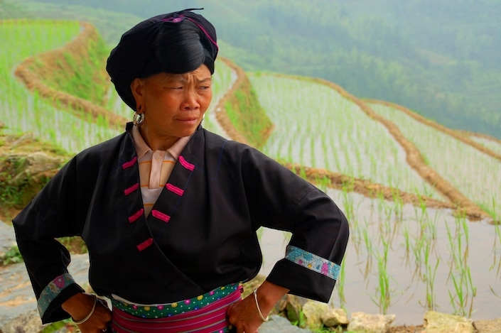 a woman in traditional Chinese ethnic minority dress stands with her hands on her hits in front of flooded rice terraces