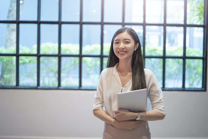 asian woman office assistance employee standing up straight smiling while holding a tablet device with both hands on her stomach, wearing a long sleeves shirt rolled up to the elbow and a pink skirt.